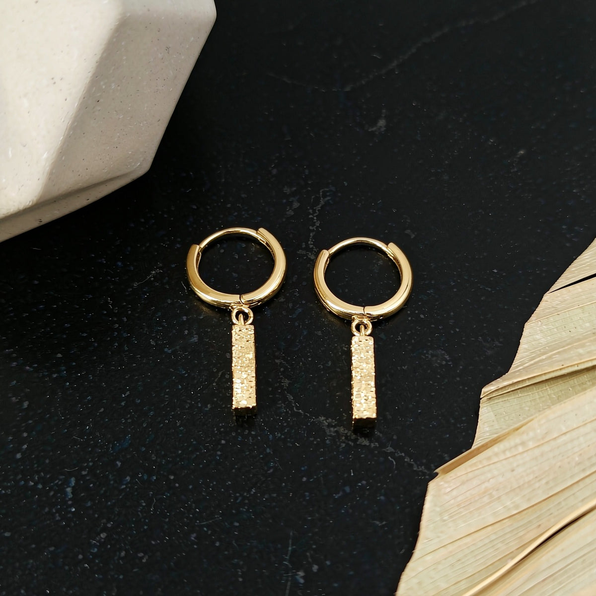 Gold Sora hammered bar drop earrings on a dark surface